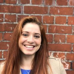 Photograph of a woman with long red hair smiling with a brick wall in the background.