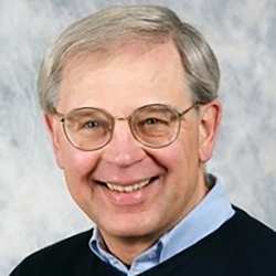 portrait of a man in a blue shirt and sweater in front of a gray wall