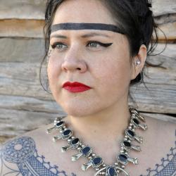 A woman with black hair and red lips looks off camera to the left. She has a black line drawn above her eyebrows across her forehead, an intricate tattoo patter from her shoulders across her collar bone, and is wearing an intricate silver and gemstone necklace.