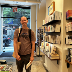Photograph of a man wearing a brown t shirt and dark slacks smiling in a small bookstore.