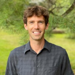 a smiling man in a gray shirt in front of a green tree and grass
