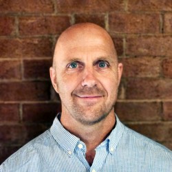 A headshot photo of David Lane, a person with green eyes and facial stubble, wearing a light blue and white pinstriped collared shirt.