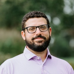 A headshot of Michael Rosato, a person with dark hair, glasses, and a beard, wearing a light pink polo shirt