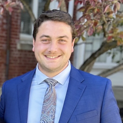 a man in a blue suit and tie smiles in front of a tree