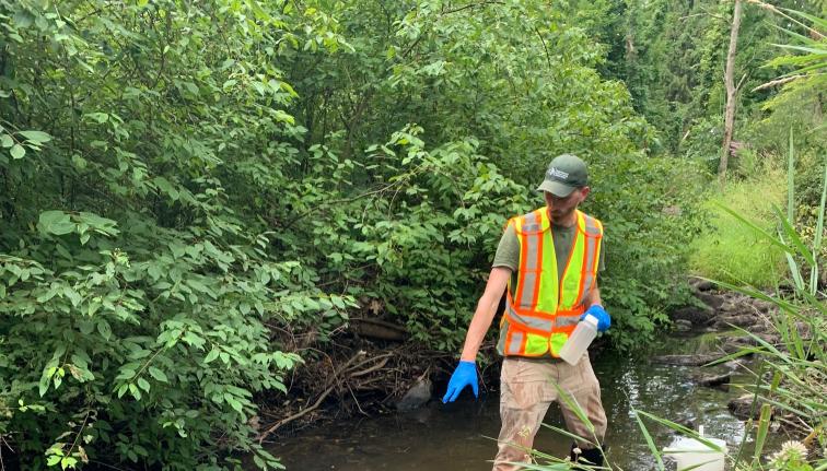 Photograph of a man wearing a bright vest, green hat, and blue rubber gloves standing in a small stream.