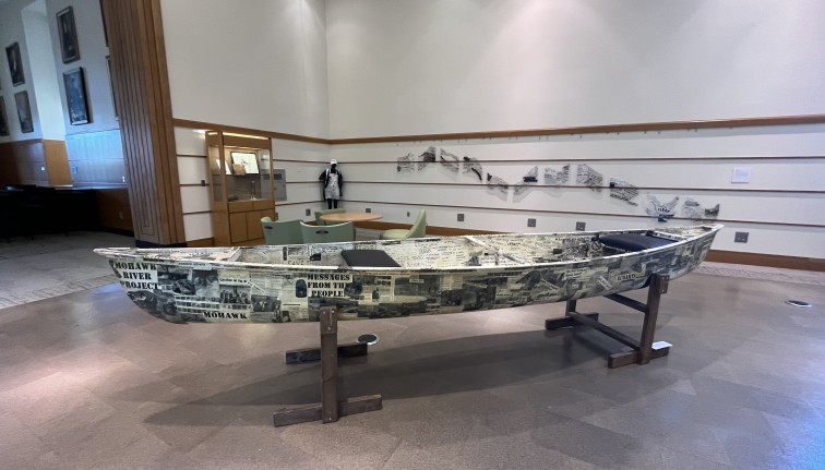 Photograph of a canoe in an exhibit covered with black and white photos and hand written messages.