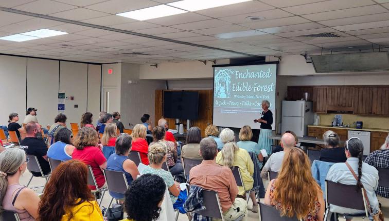 A room full of people seated and facing a presenter who is standing in front of a projector screen with a slide on it.