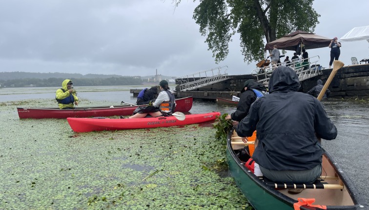 Photograph of two canoes and one kayak with five people on them in the middle of a river filled with green vegetation.