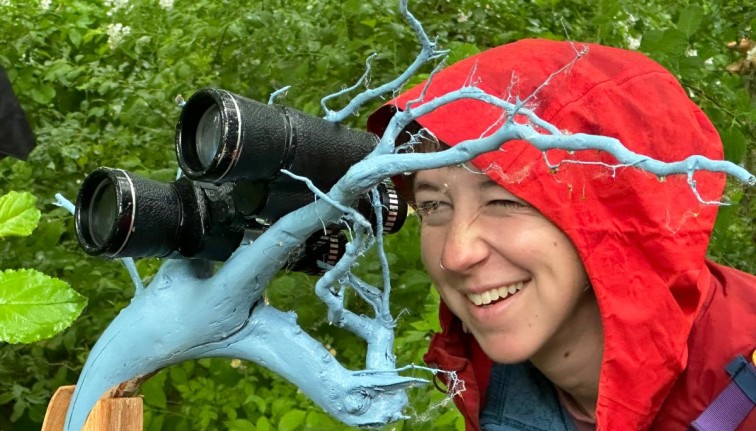 Photograph of a person wearing a rain coat with the hood on their head looking through a pair of binoculars on a blue painted branch.