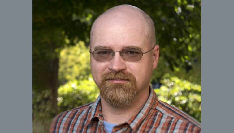 A headshot of a person with glasses and a light-colored goatee standing in front of a sun-dappled grove