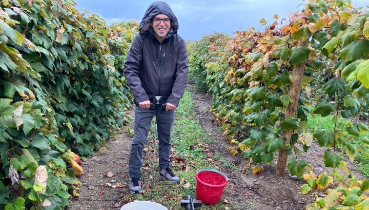 A person in a raincoat stands facing the camera, holding a soil core sampler tool in a vineyard in mid-spring. A white bucket in front of them is ready to collect the sample.