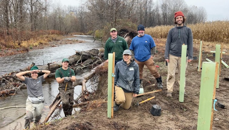 Photograph of a group of people standing on a creek shore with freshly planted trees in tree tubs.