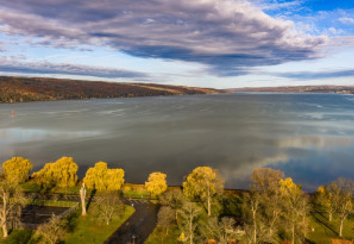 A wide aerial view of a calm lake surrounded by rolling hills in autumn colors. Golden trees line the shoreline in the foreground, and the sky above is filled with dramatic clouds reflecting on the water’s surface.