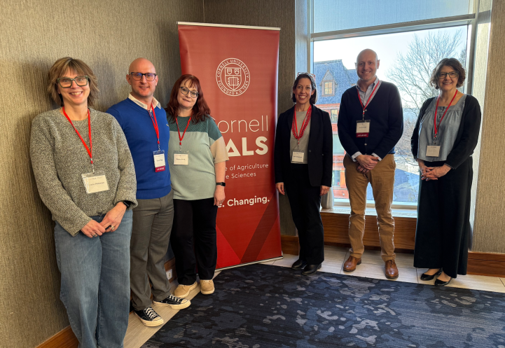 6 individuals stand and smile in front of Cornell CALS poster