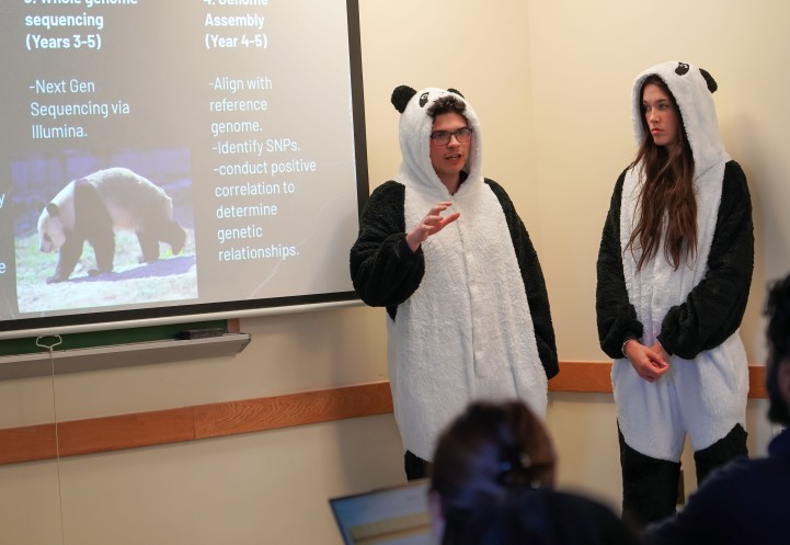a man and a woman dressed in panda onsies stand by slide projection on the genetics of pandas