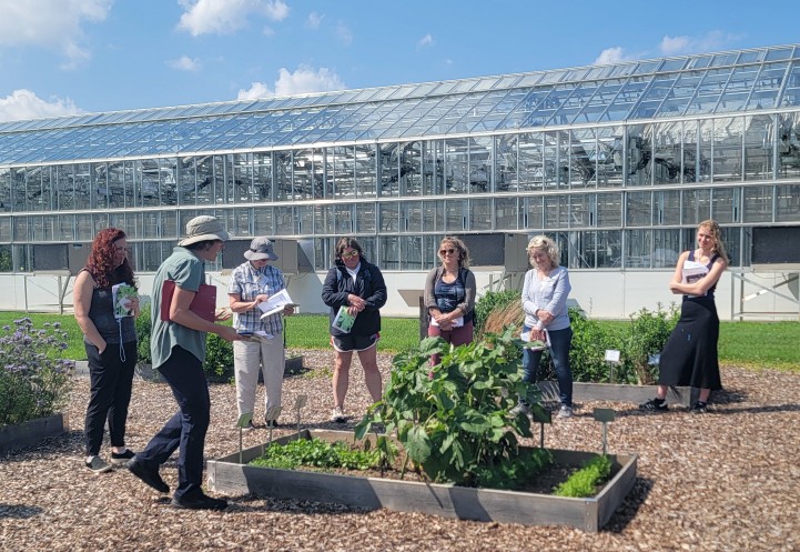 A person stands before a group, infront of a raised garden bed, with green houses in the background. 