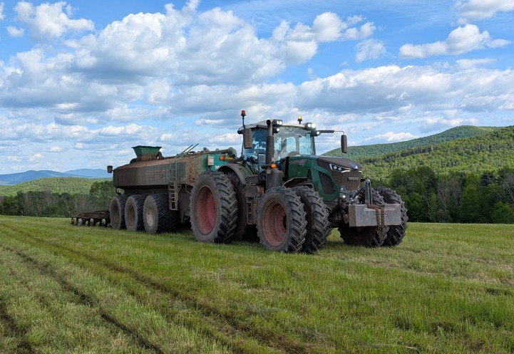 A large green tractor with a manure spreader stands in a green field, surrounded by rolling hills and a partly cloudy sky.