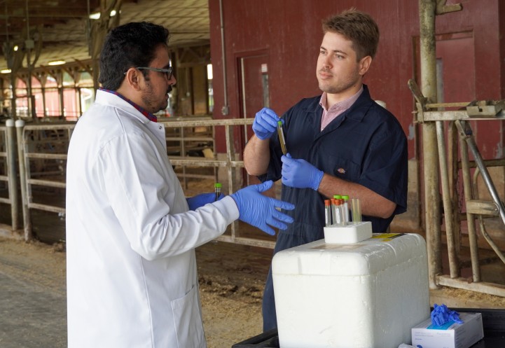 a man holds a vial of liquid and talks with another man in a white lab coat in a barn