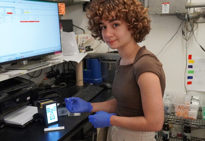 a woman wearing rubber gloves holds a plastic cassette is about to put it into a small square piece of tech with a touch screen