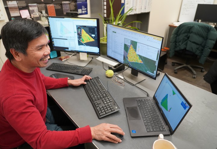 A man works on three computers. He is wearing a red shirt, sitting at a desk and smiling.