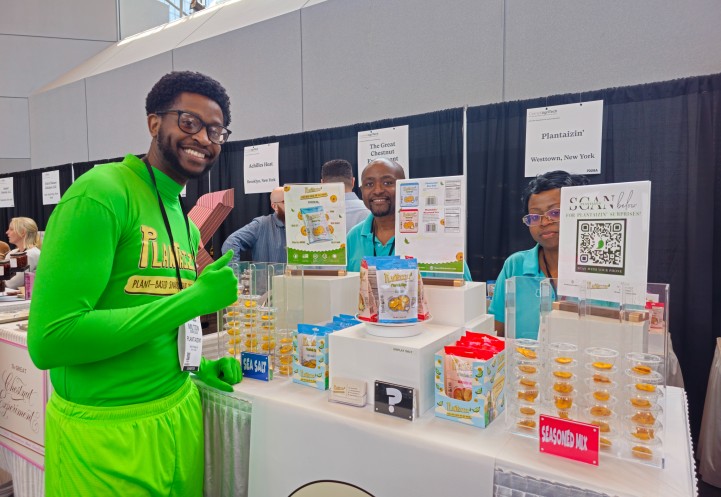 Three people, one of whom is wearing a green body suit, standing at a trade show booth. 