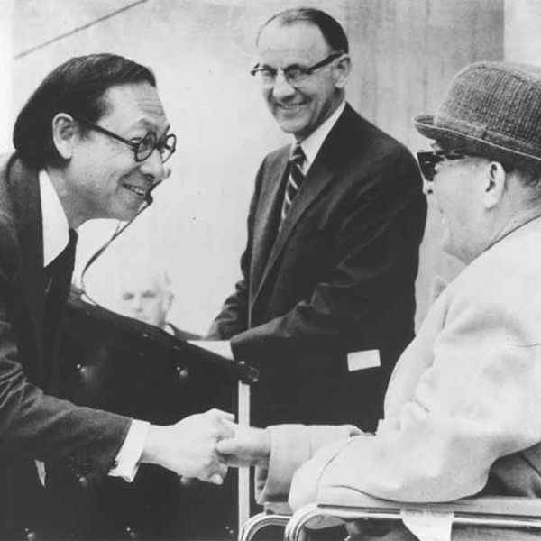 2 men shaking hands with one man in background