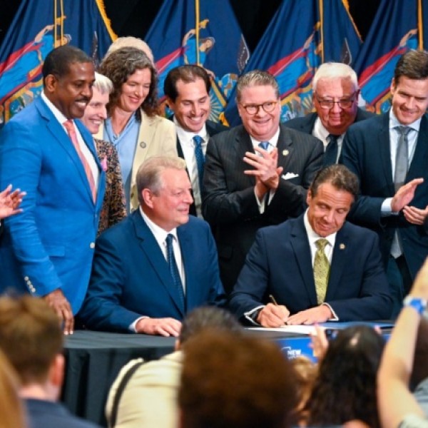 In a press room, a man in a suit signs a bill while others stand behind him watching and clapping