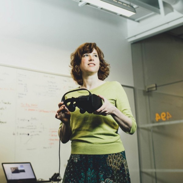 A woman standing in a classroom looking off to the side while holding a pair of virtual reality goggles