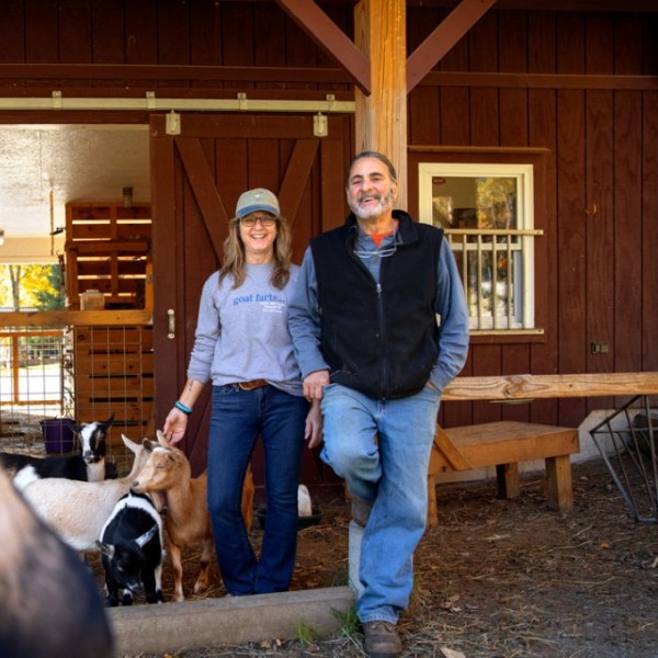 A man and woman standing with their back to a barn with small goats at their feed