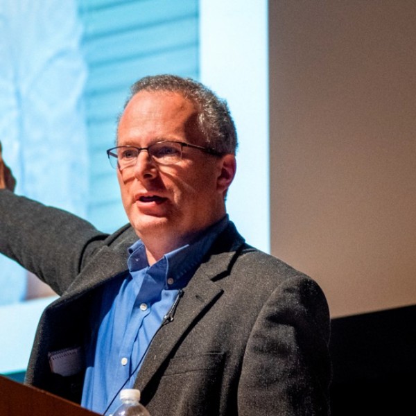 A caucasian man standing at a podium talking and pointing to the screen behind him