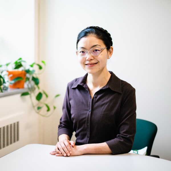 An asian female sitting at a table with her hands crossed. She is wearing glasses and a purple button down shirt and has her hair tied up in a braid. She is sitting in a white-walled office.