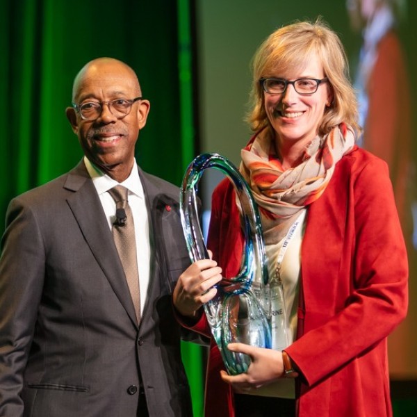 A man and woman standing on a stage smiling while the woman holds a large class award