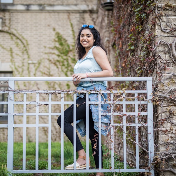 A female student standing and smiling standing behind a fence in front of a tan building