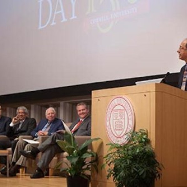 Five people sit on chairs on stage while one man stands at a podium and speaks