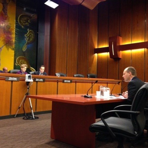 One man sits and faces a group of people sitting in front of the room and facing him