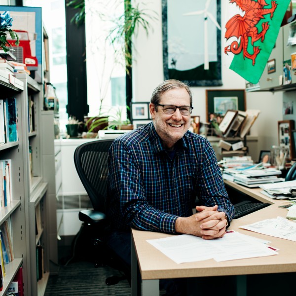 A man poses happily in an office while sitting with hands clamped on a desk