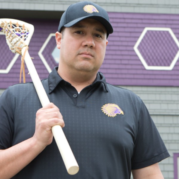 Picture of man holding a lacrosse stick in front of the side of a grey and purple building.