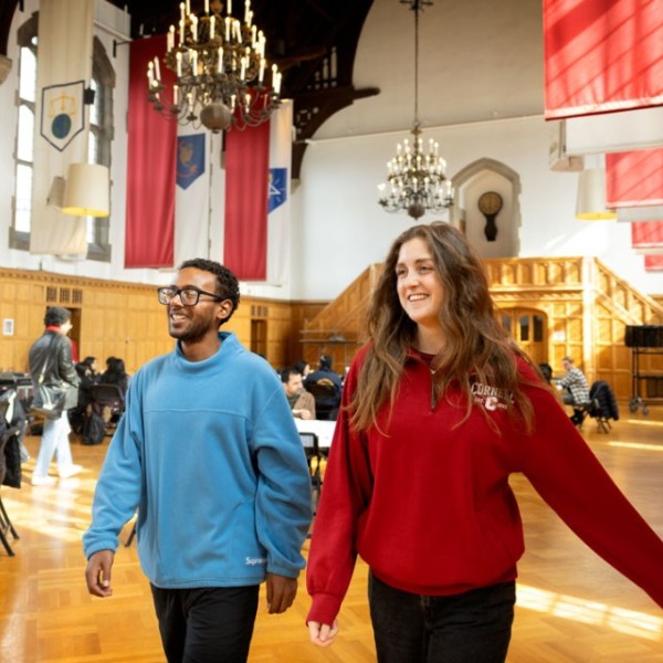 A man in a blue crewneck and a woman in a red cornell quarter zip smile while walking in the willard straight hall memorial room.