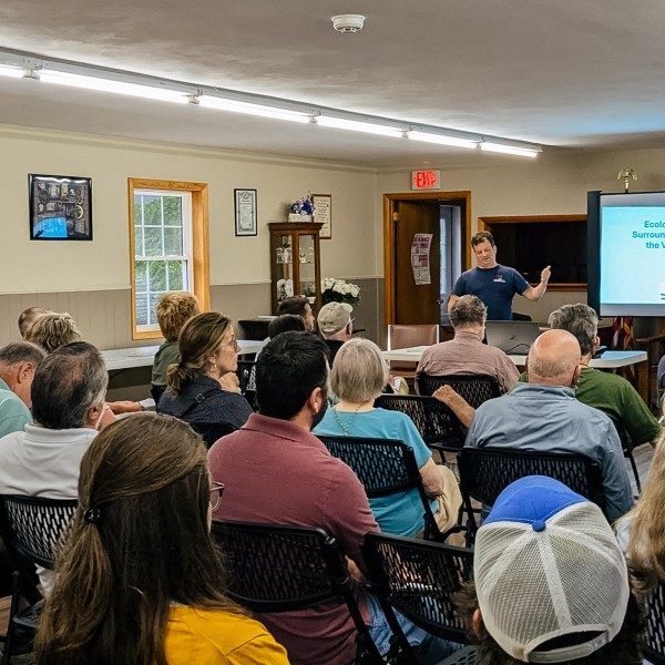 Photograph of a man standing at the front of a room with slides projected talking to about twenty people seated in rows.