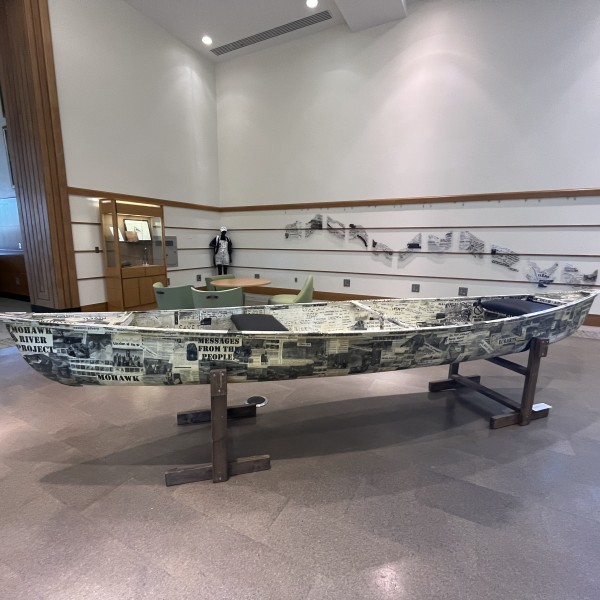 Photograph of a canoe in an exhibit covered with black and white photos and hand written messages.