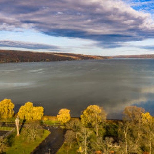 A wide aerial view of a calm lake surrounded by rolling hills in autumn colors. Golden trees line the shoreline in the foreground, and the sky above is filled with dramatic clouds reflecting on the water’s surface.