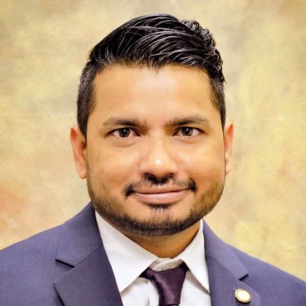 A headshot photo of Kiran Gadhave, wearing a blue suit, pastel shirt and navy blue tie, and smiling at the camera.