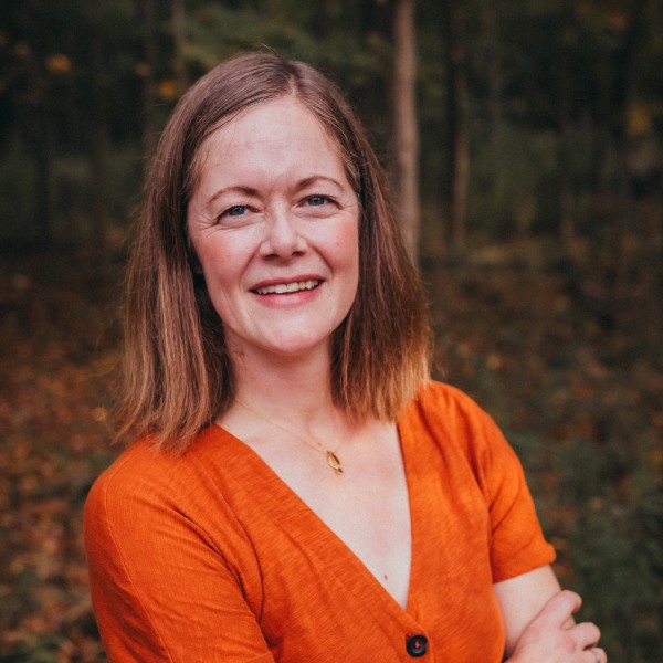 Professional portrait of person standing with crossed arms and long brown hair wearing an orange shirt.