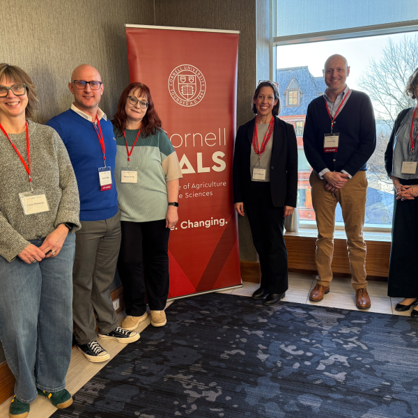 6 individuals stand and smile in front of Cornell CALS poster