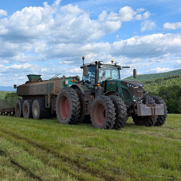 A large green tractor with a manure spreader stands in a green field, surrounded by rolling hills and a partly cloudy sky.