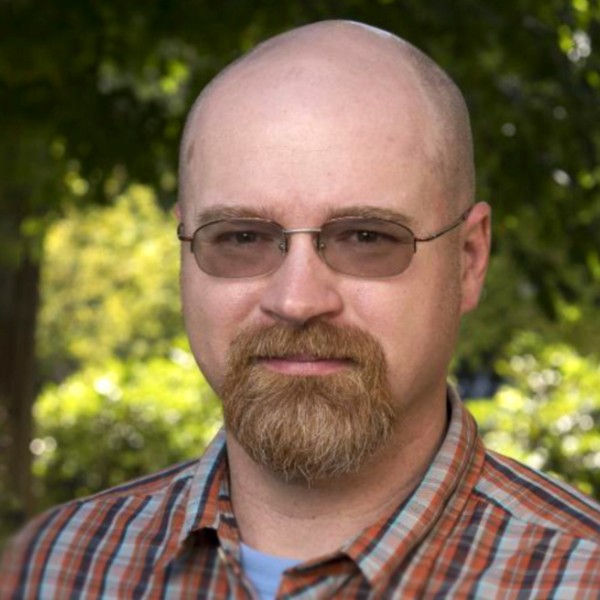 A headshot of a person with glasses and a light-colored goatee standing in front of a sun-dappled grove