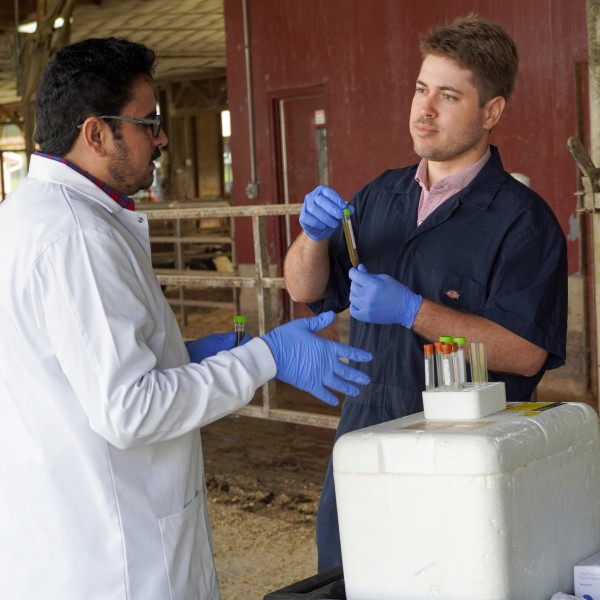 a man holds a vial of liquid and talks with another man in a white lab coat in a barn
