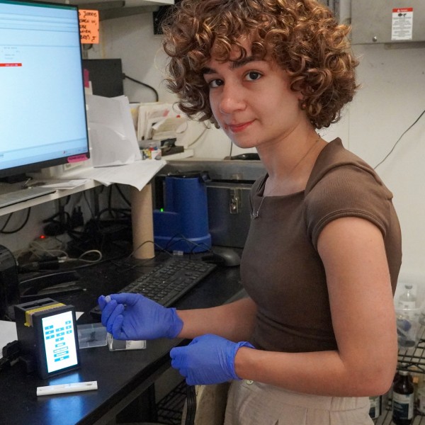 a woman wearing rubber gloves holds a plastic cassette is about to put it into a small square piece of tech with a touch screen
