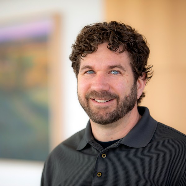 A head-and-shoulders portrait of a smiling man looking directly at the camera. He has curly, dark brown hair, a full beard and mustache, and bright blue eyes. He is wearing a dark gray, short-sleeved polo shirt with a fine, horizontally ribbed texture. The background is blurred, showing a framed artwork with soft, abstract colors on the left and a plain, light wood-toned surface on the right.