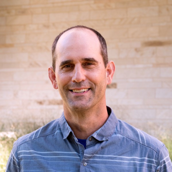 A portrait of a middle-aged man smiling directly at the camera. He has short dark hair on the sides of his head, and light stubble. He is wearing a blue button-down shirt with thin horizontal white stripes. The background is an out-of-focus light tan stone wall with some blurred green vegetation at the bottom.